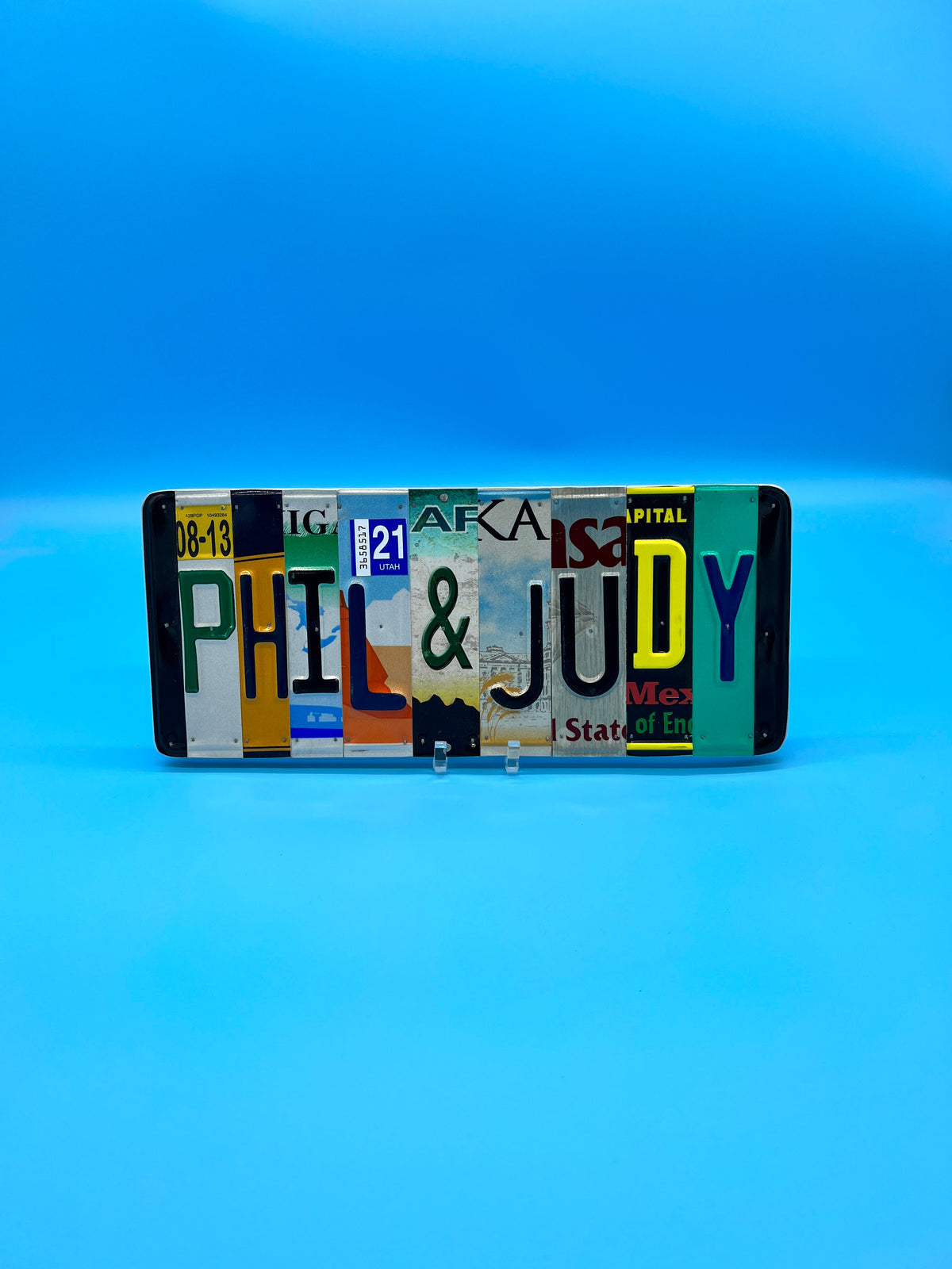 A license plate sign made from multiple colorful license plates arranged to spell out 'PHIL & JUDY', displayed against a blue gradient background.