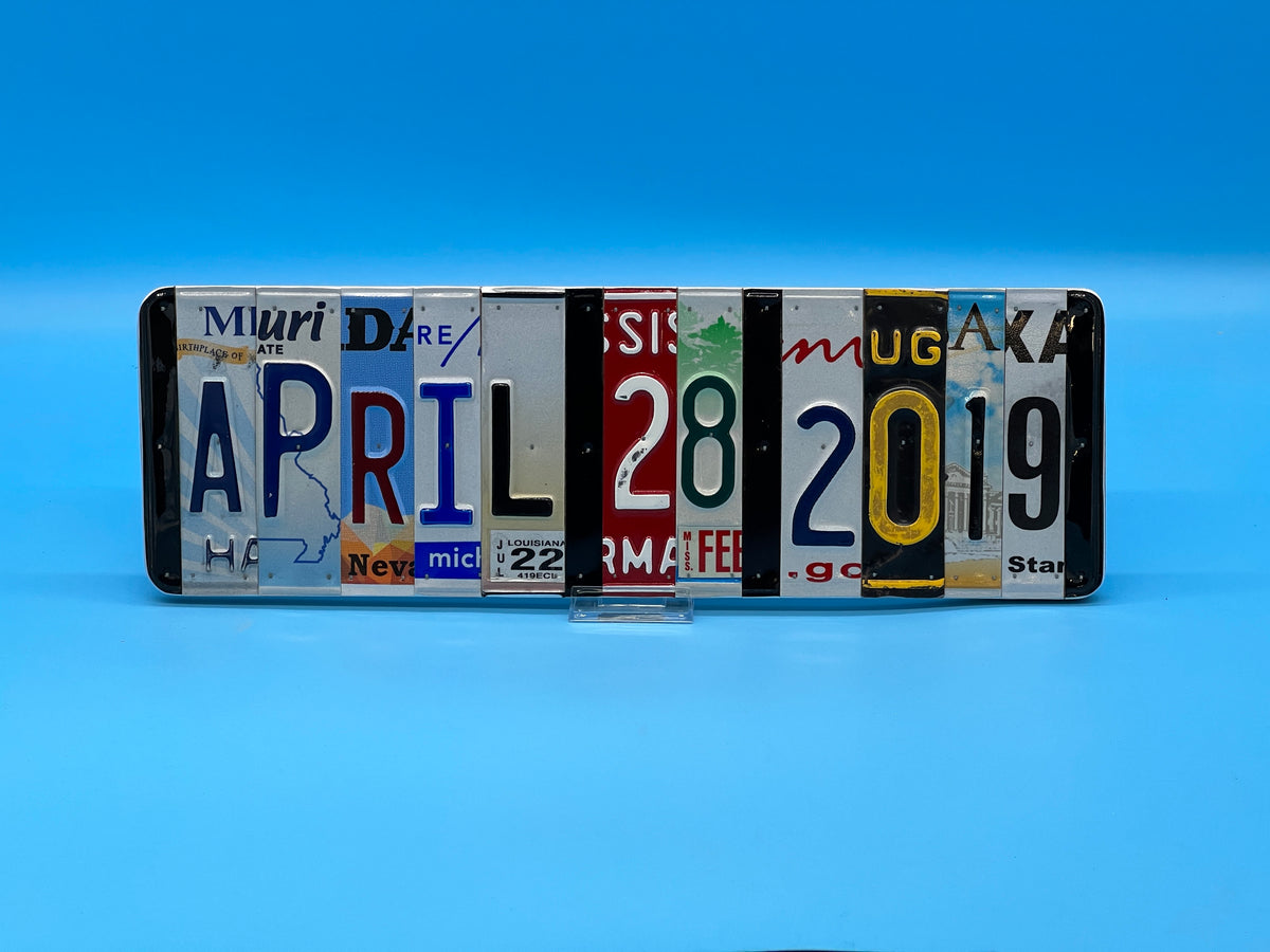A rectangular decorative wall piece made from seven vintage license plates arranged side by side to spell out April 28, 2019, displayed on a small clear stand against a blue gradient background.