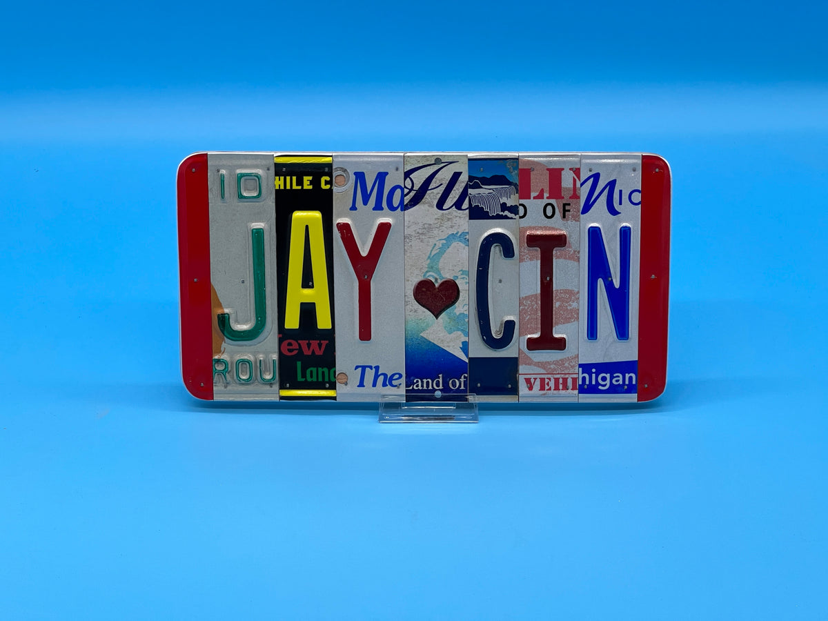 A rectangular novelty sign made from cut-up license plates spelling 'JAY CIN' with a small heart in the center, displayed on a solid blue background.