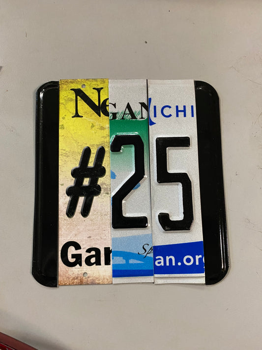A square sign made with license plate from Michigan and New Jersey cutup and nailed to wood with rounded corners.  The number 25 in black over a multicolored background of white, blue, green, and yellow.