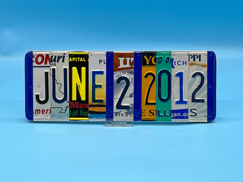 A license plate sign made from six license plate letters and numbers arranged vertically to spell out the date "June 2012," standing on a small, clear display block against a blue gradient background.