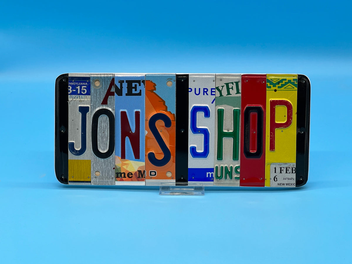 A sign made from colorful vintage license plates spelling out 'JON'S SHOP', displayed on a clear stand against a blue gradient background.