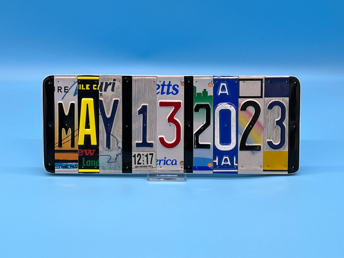 A novelty license plate sign displaying the date May 13, 2023, made from different license plates mounted on wood. Hanging against a solid blue background.