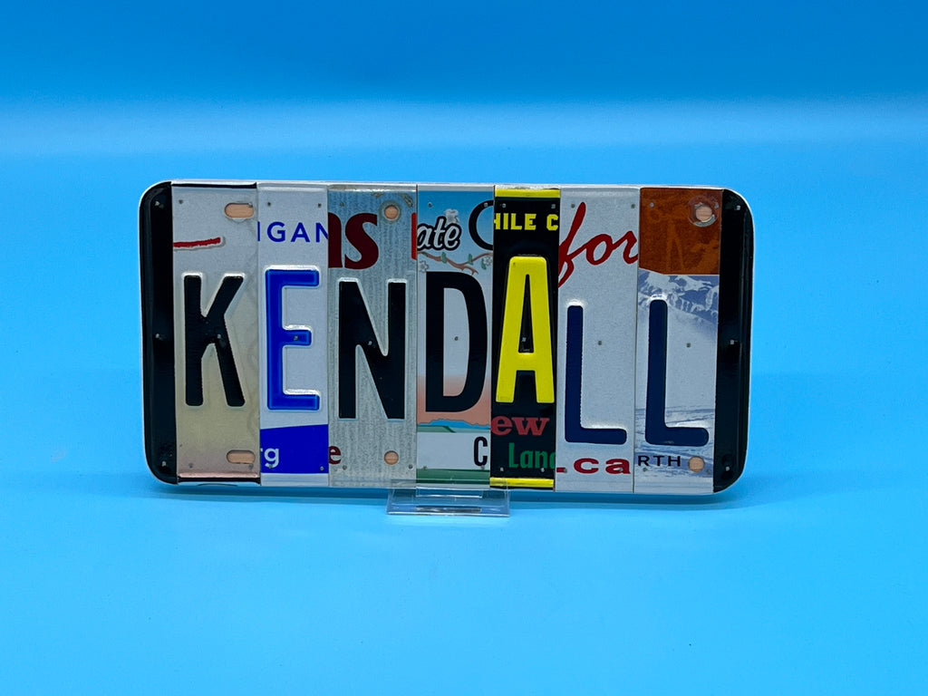 A custom wall sign spelling 'KENDALL' in raised letters, crafted from various license plates, displayed on a solid blue background.