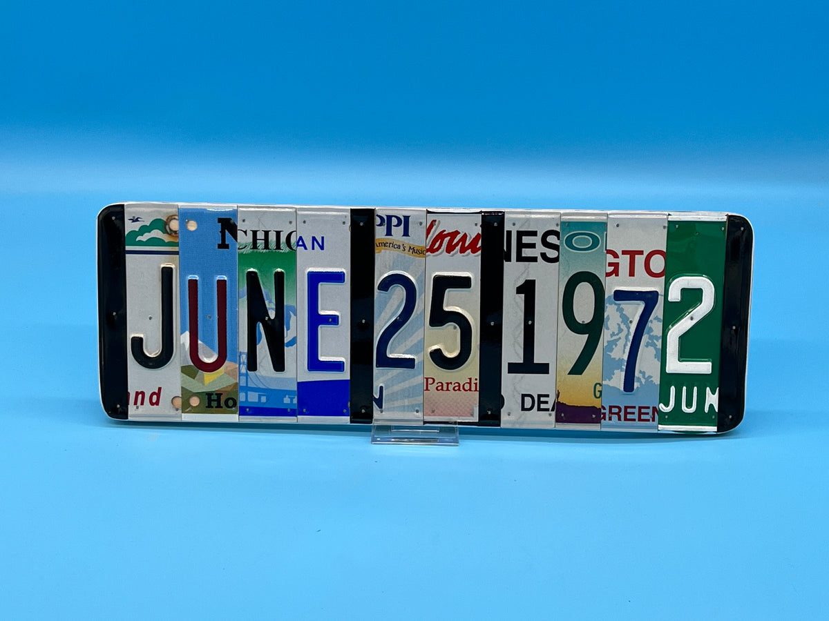 A novelty wall sign made from cut-up vintage license plates spelling out 'JUNE 25 1972', displayed on a small clear stand against a blue gradient background.
