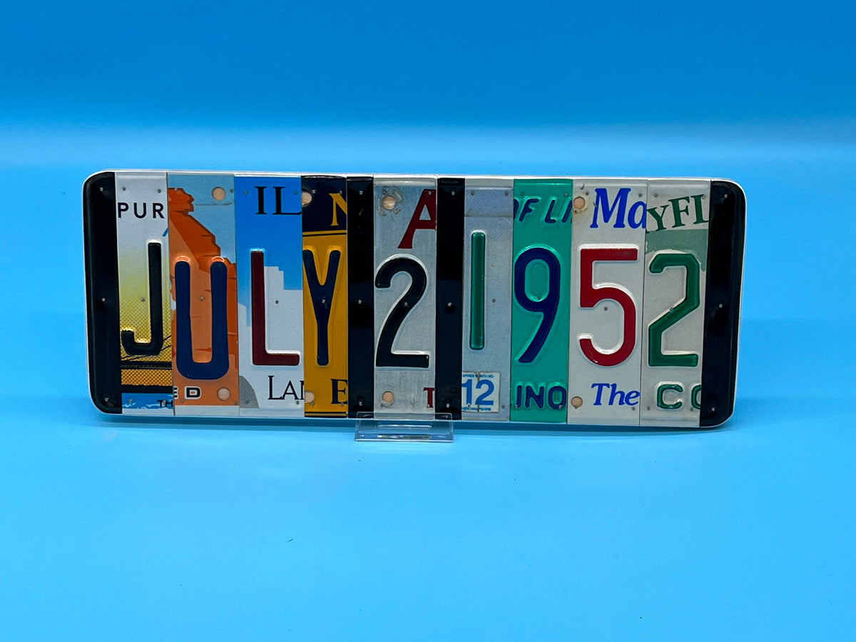 A novelty sign made from a row of cut-up license plates spelling out 'JULY 21 1952'. Against a solid blue background.
