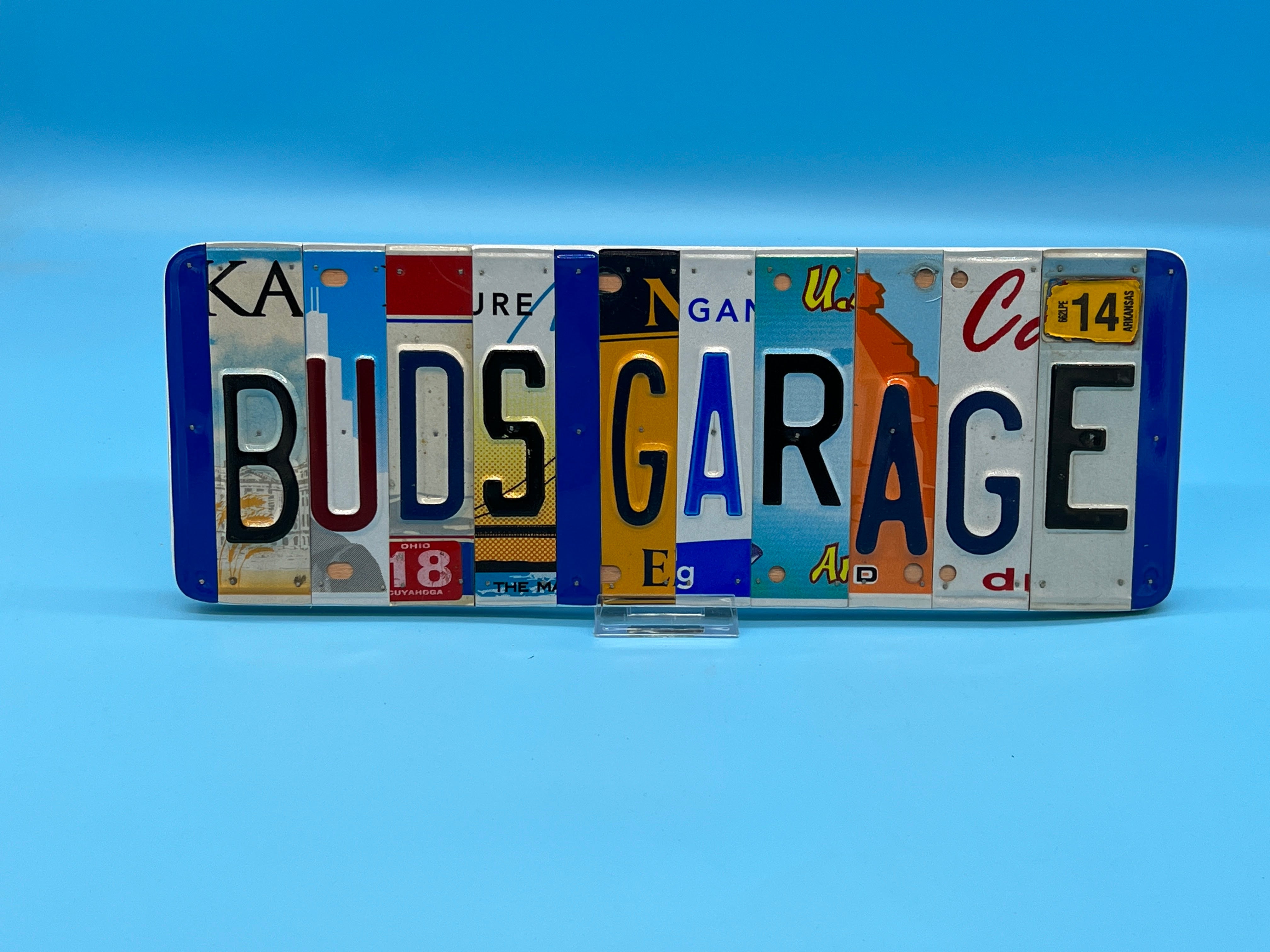 A rectangular decorative sign spelling 'BUDS GARAGE' made from multiple vintage license plates, displayed on a small clear stand against a solid blue background.