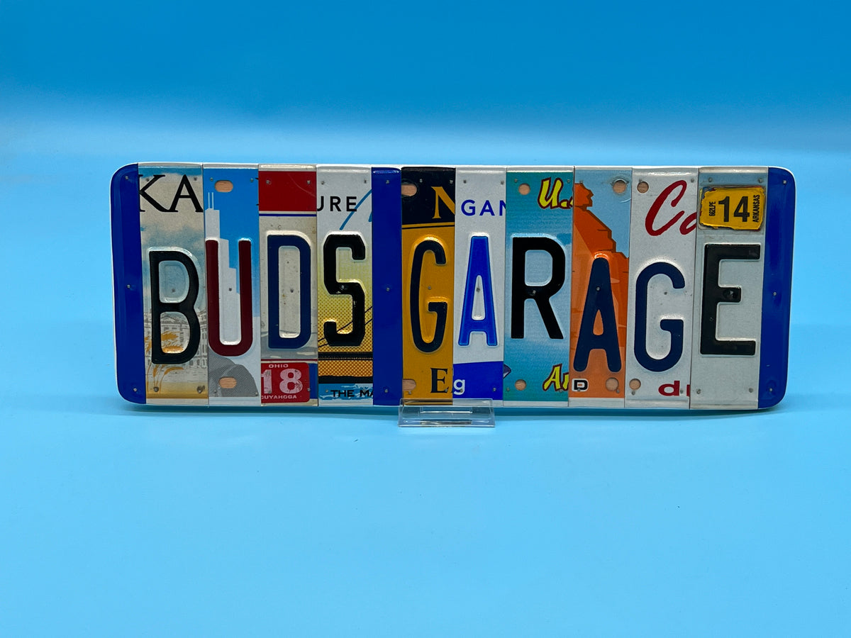 A rectangular decorative sign spelling 'BUDS GARAGE' made from multiple vintage license plates, displayed on a small clear stand against a solid blue background.