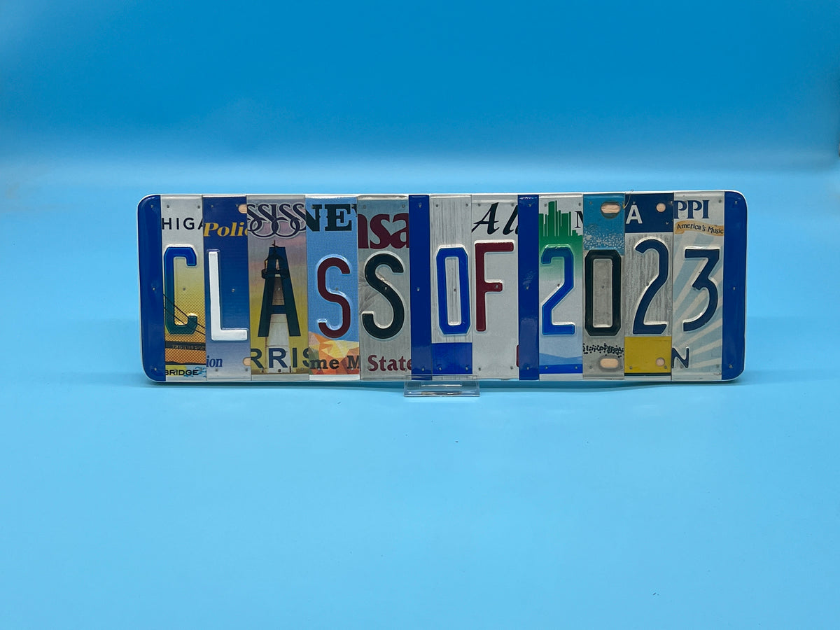 A novelty license plate sign spelling out 'CLASS OF 2023' using various colorful license plates, displayed on a small clear stand against a blue gradient background.