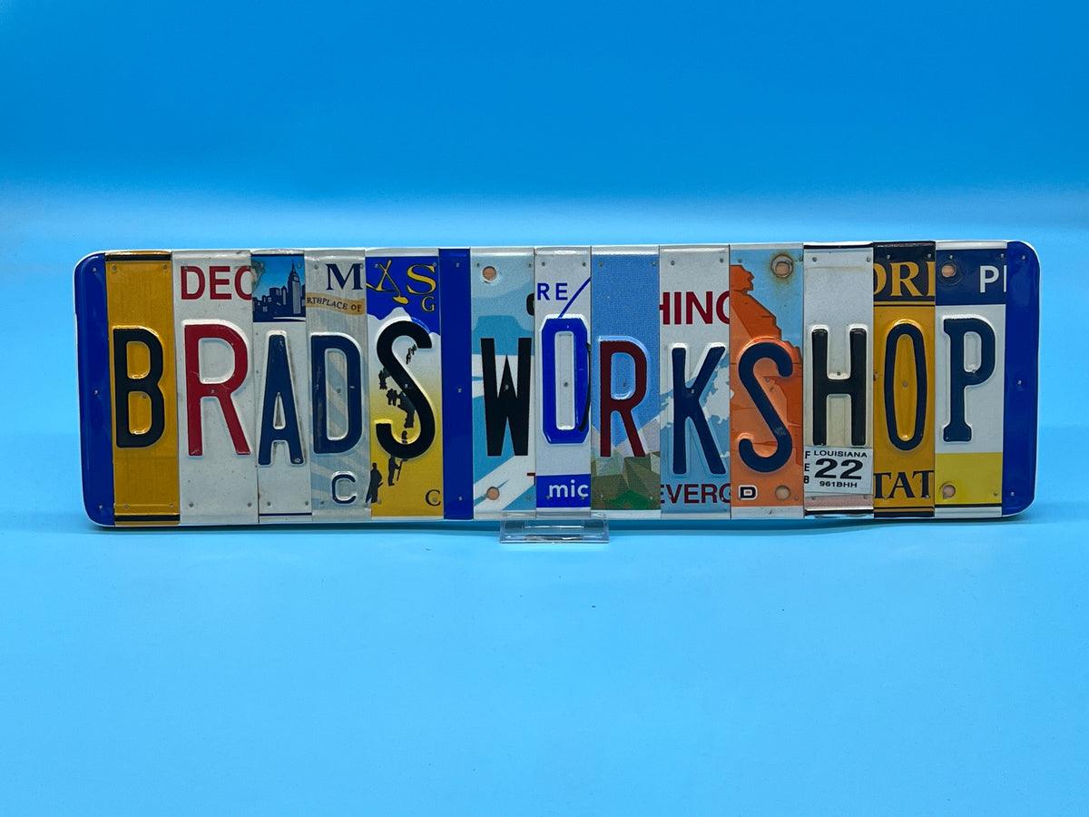 A decorative license plate wall sign spelling 'BRADS WORKSHOP' with various colorful vintage license plates, displayed on a clear stand against a blue gradient background.