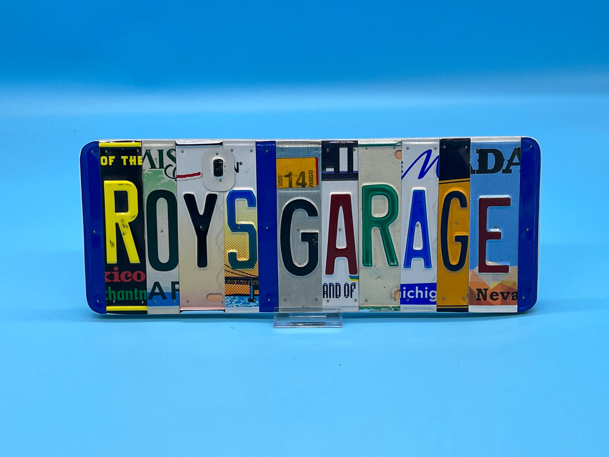 A rectangular phone case made from cut-up vintage license plates spelling 'ROYS GARAGE', displayed on a clear stand against a blue gradient background.
