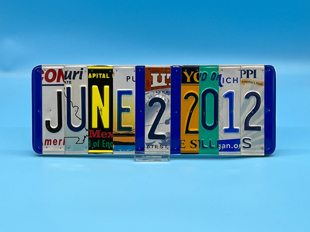 A novelty wall sign made from recycled license plates cut vertically and nailed to wood. Spelled out the date June 2 2012, standing on a small clear display block against a blue gradient background