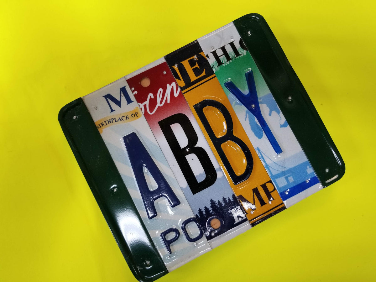 License plate letters from different states nailed to wood, the name ABBY. With a bright yellow background