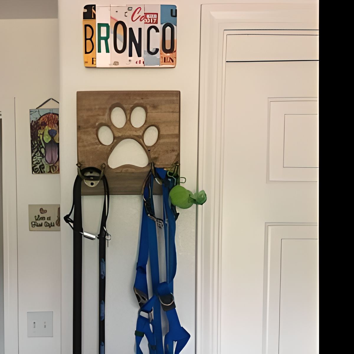 A wooden paw-print shaped wall hook rack hanging on a white doorframe, holding dog leashes and a blue harness, with a decorative license plate sign reading BRONCO above.