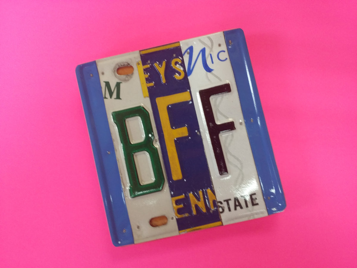 A square license plate sign featuring a colorful collage of vintage license plate letters BFF set against a solid hot pink backdrop.