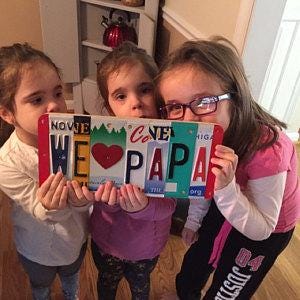 Three children holding up a sign made of license plates with 'We love Papa' text.                                                                                      