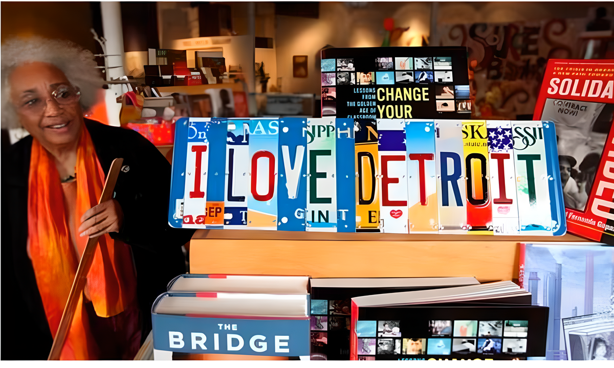 A woman with white hair and glasses, wearing a bright orange scarf, stands smiling next to a display of bookshelves and a colorful license plate sign spelling 'I LOVE DETROIT' in a bookstore.