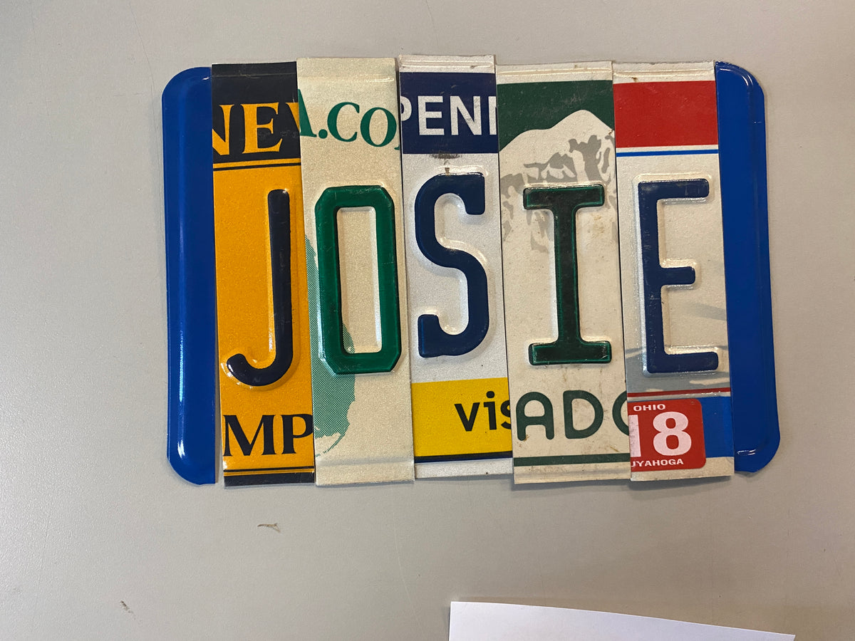 A handmade license plate wall sign spelling 'JOSIE' using various vintage license plates with blue borders, set against a plain light gray wall.