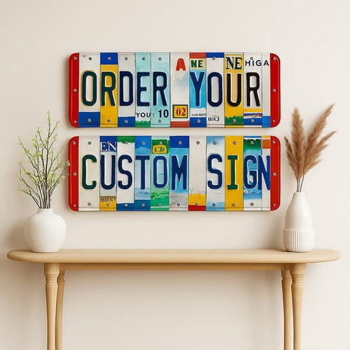 Two rectangular license plate wall signs mounted on a beige wall above a light wood console table. The top sign reads “ORDER YOUR” and the bottom sign reads “CUSTOM SIGN,” each made from colorful license plate strips with red, white, blue, yellow, green, and black lettering.