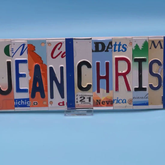 A custom wall sign spelling 'JEAN CHRIS' in raised letters, crafted from various license plates, displayed on a solid blue background.