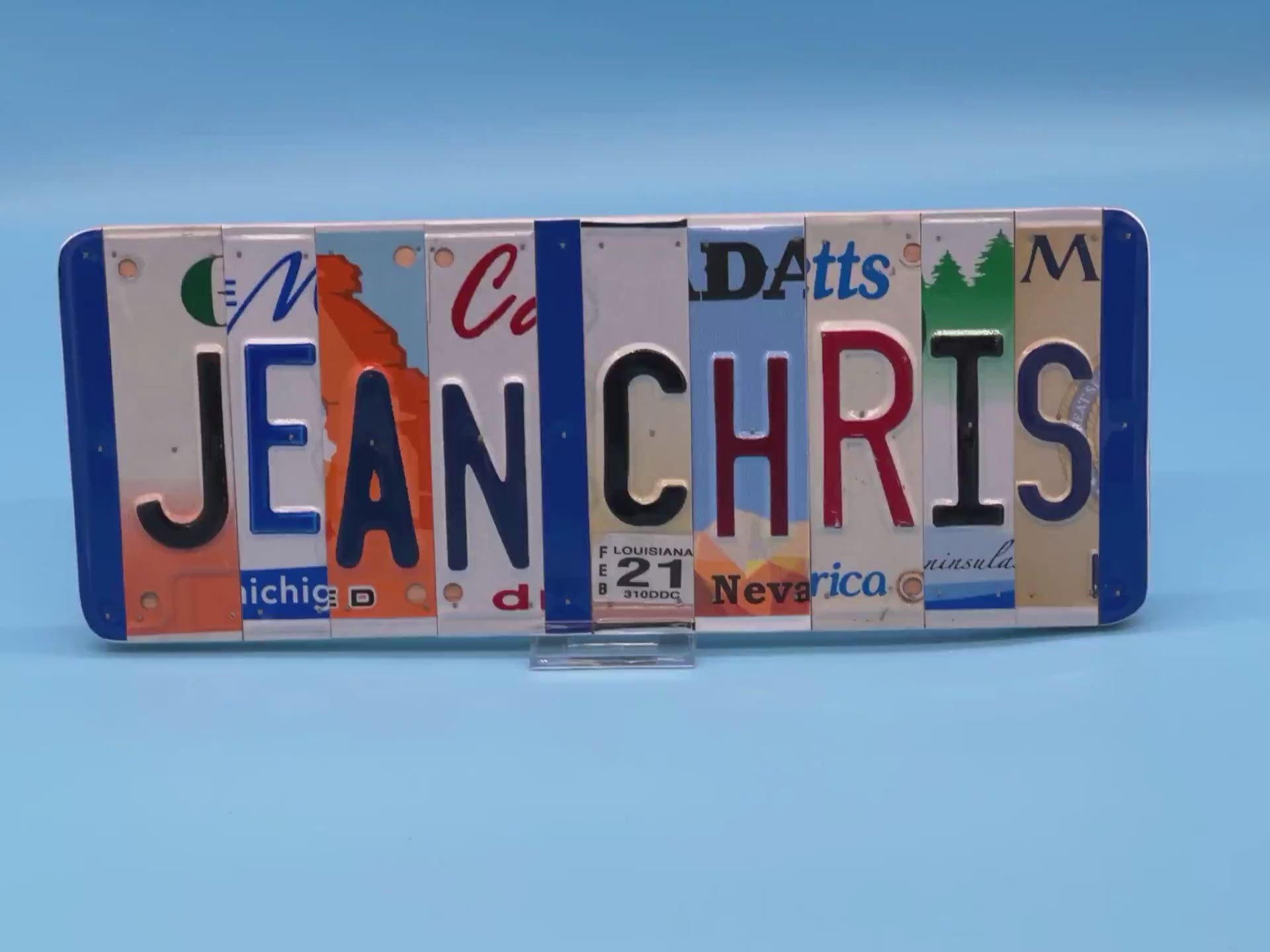 A custom wall sign spelling 'JEAN CHRIS' in raised letters, crafted from various license plates, displayed on a solid blue background.