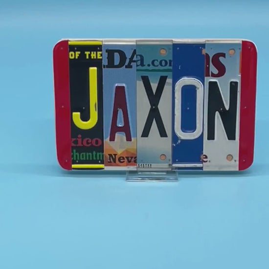 A custom wall sign spelling 'JAXON' in raised letters, crafted from various license plates, displayed on a solid blue background.