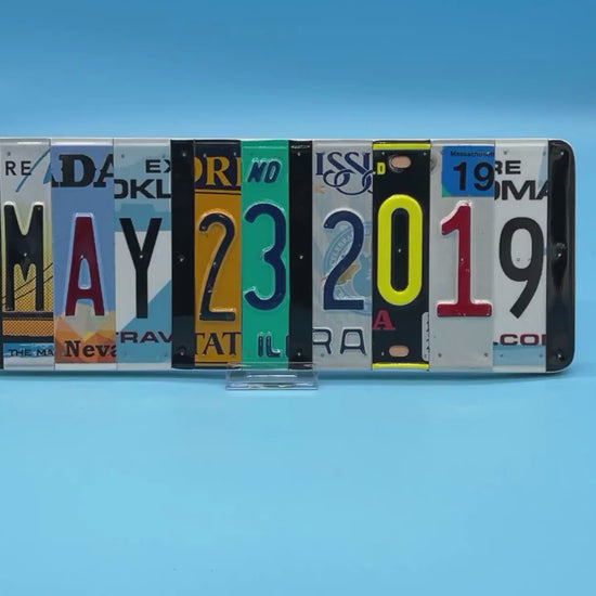 A rectangular decorative wall piece made from cut-up license plates arranged side by side to spell out MAY 23, 2019.