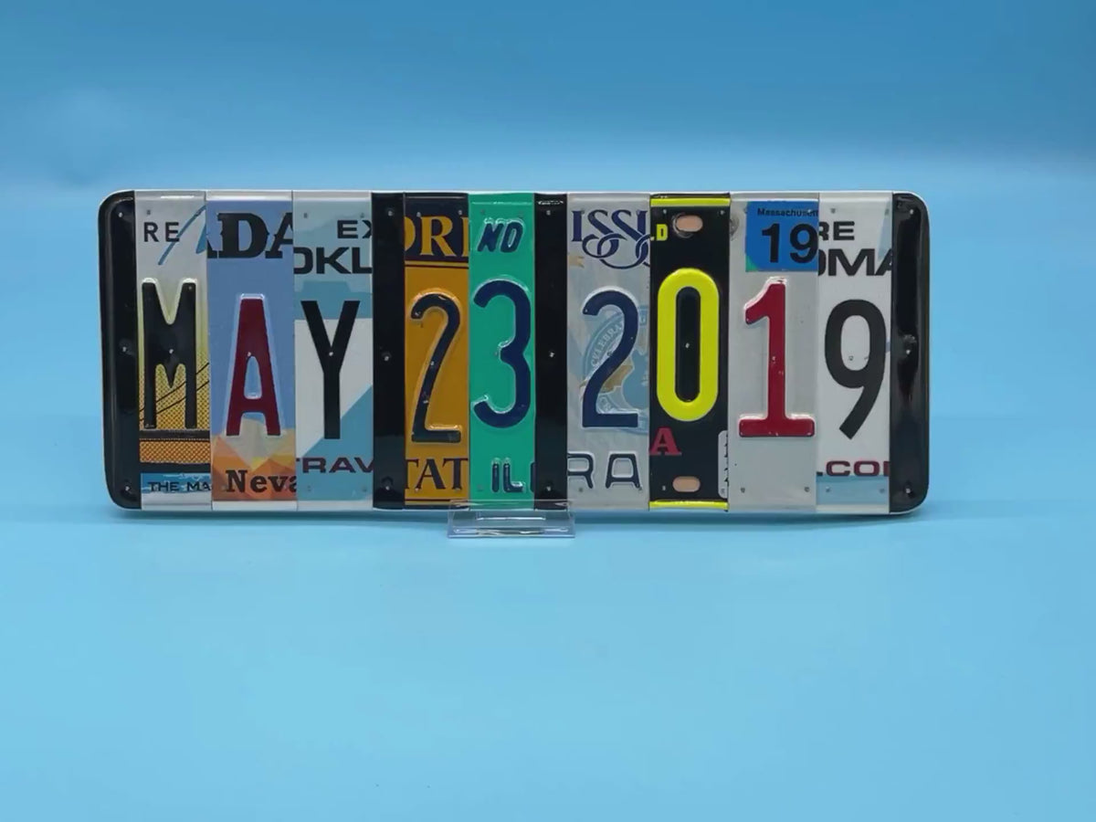 A rectangular decorative wall piece made from cut-up license plates arranged side by side to spell out MAY 23, 2019.