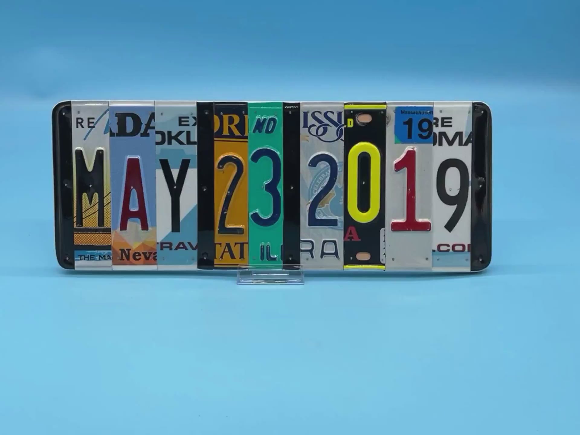 A rectangular decorative wall piece made from cut-up license plates arranged side by side to spell out MAY 23, 2019.